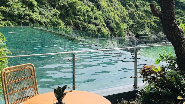 A wooden deck with a round table and wicker chair overlooking a clear water body with a forested hillside in the background. - Forest Sky Villa - Flamingo Cat Ba (Hai Phong)