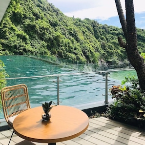 A wooden deck with a round table and wicker chair overlooking a clear water body with a forested hillside in the background.