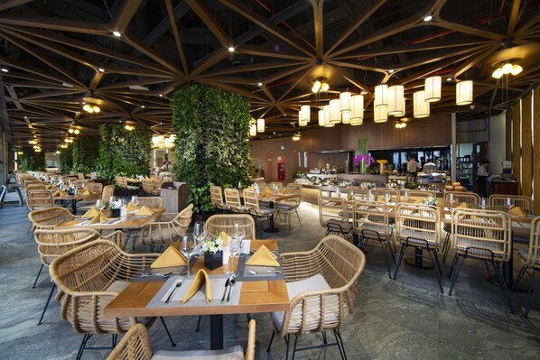 A spacious restaurant with high ceilings, wicker furniture, and a central green wall.