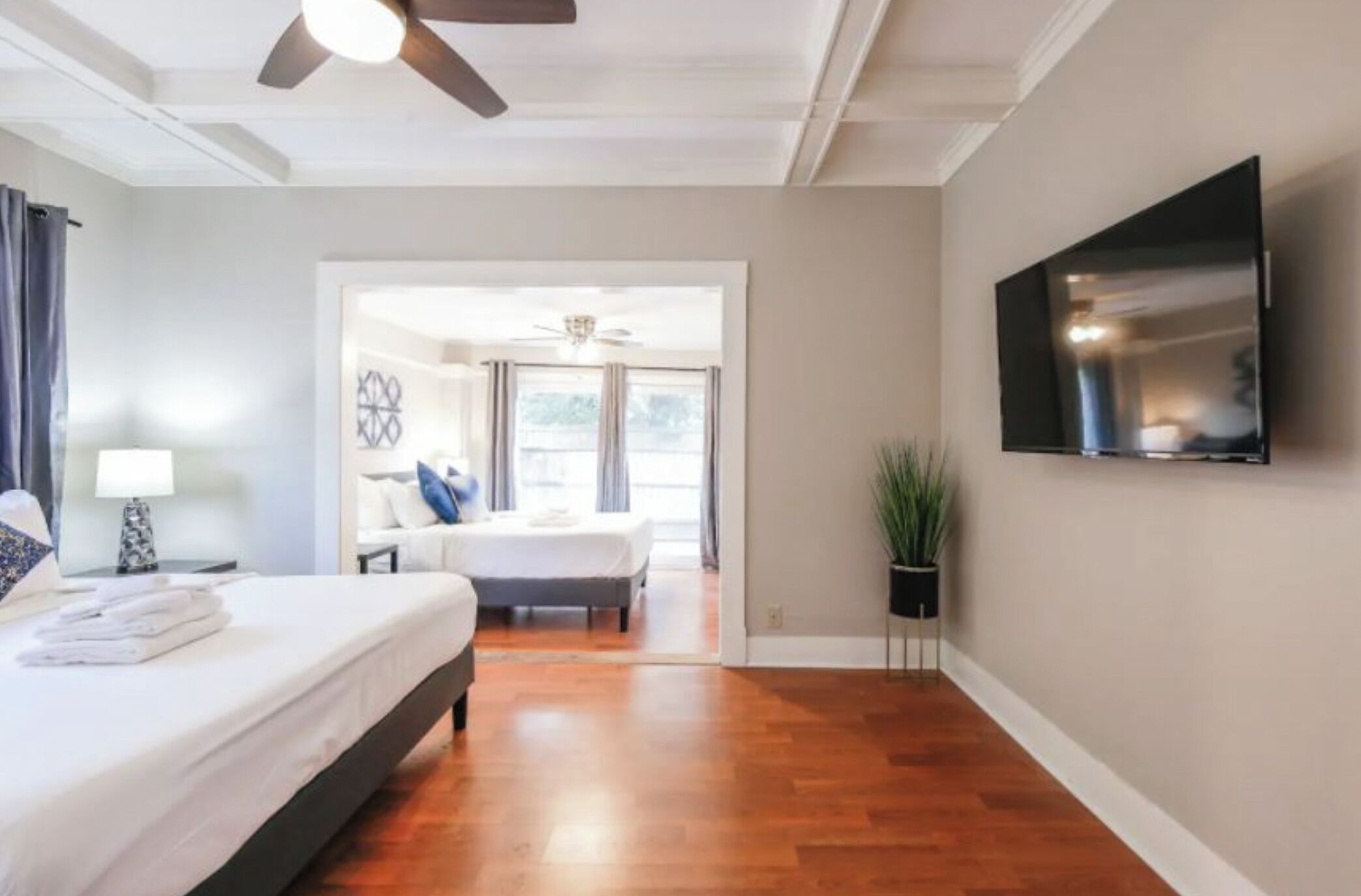 Habitación de hotel con cama, televisión montada en la pared, ventilador de techo y vista hacia la habitación contigua.
