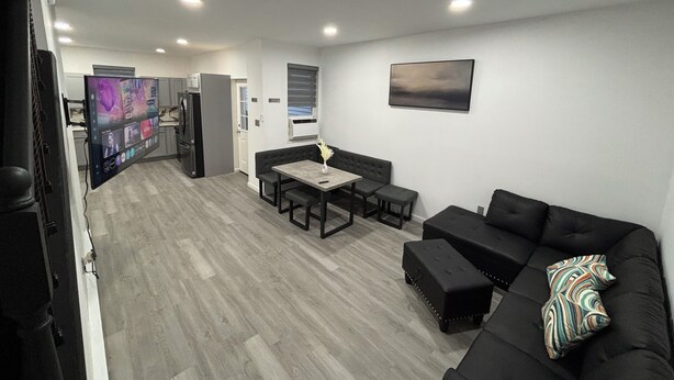 A modern living room with a sofa, a dining table with chairs, a flat-screen TV mounted on the wall, and a kitchen area in the background. - 4 Beds • Parking • Netflix • W/D • Fast Wifi (Wilkes-Barre)