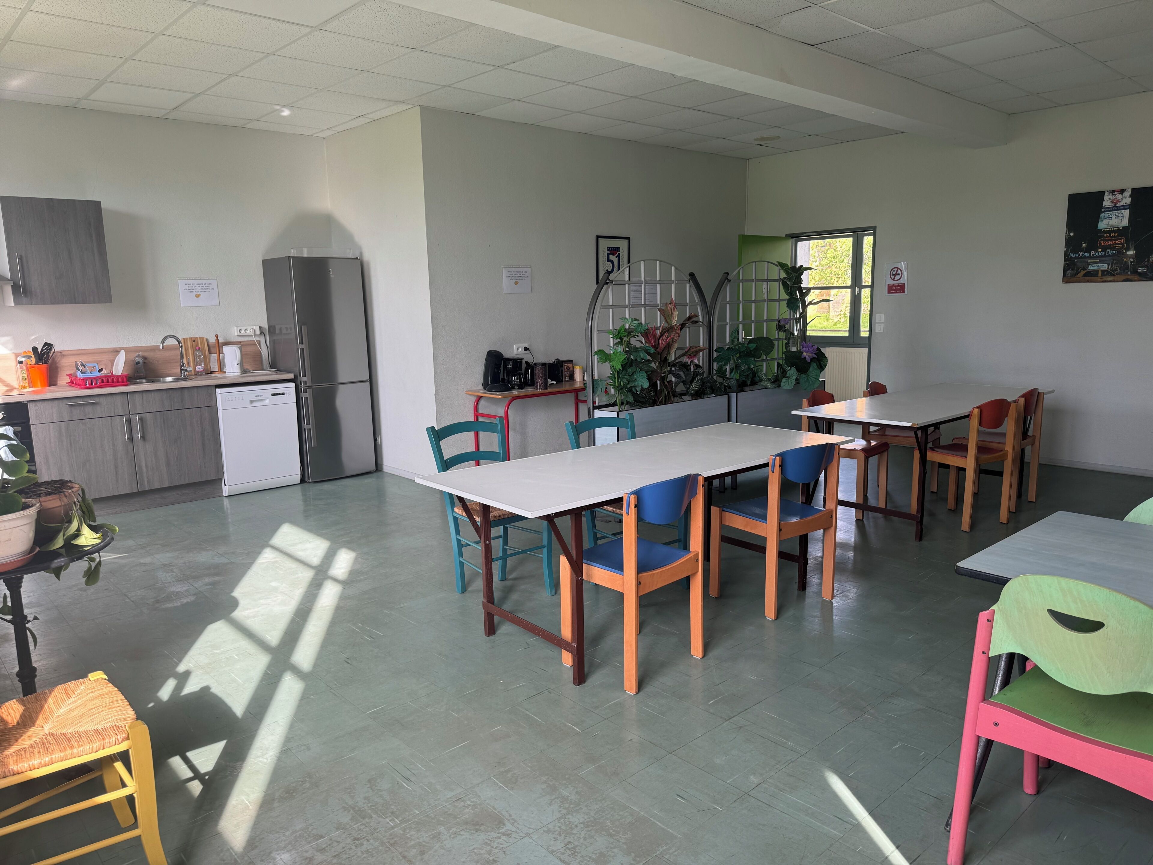 Een eethoek met tafels en stoelen, een keuken met kasten en apparatuur, en een raam met planten.
