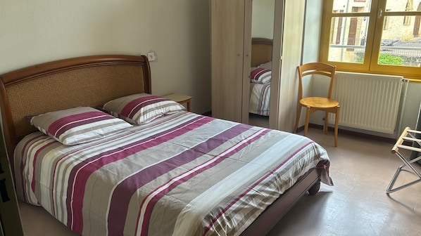 Una cama con colcha a rayas, una silla de madera, un radiador y una ventana con vistas a edificios.
