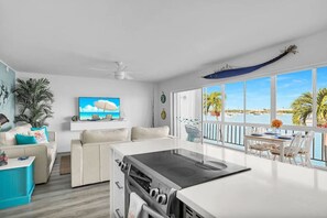A modern kitchen with a large island, a dining area with a view, and a living room with a TV and plant. - Oceanview Condo w/ Pool + Direct Beach Access (Marathon)