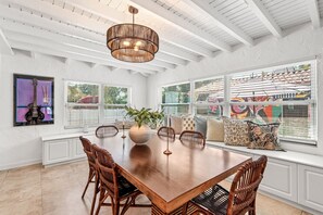 A dining area with a large wooden table, wicker chairs, a vase with a plant, and a hanging light fixture. - Spanish-Style Modern Home | Heated Pool + Hot Tub (Miami)
