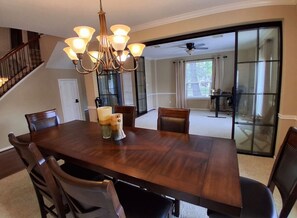 Un comedor con una gran mesa de madera, seis sillas, una araña y vistas a una sala de estar con puerta corredera de vidrio.