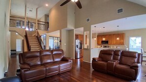 A spacious living area with a high ceiling, a staircase, leather sofas, and a kitchen area. - Lake Houston Retreat | Walk to Lake + Golf | 4BR (Humble)