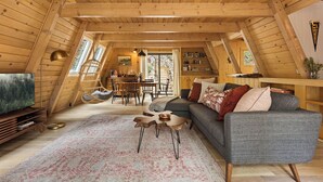 Ein gemütliches Lofts-Zimmer mit Holzbalkendecke, einem grauen Sofa, einem Esstisch mit Stühlen und einem Fernseher.