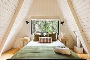 Ein Schlafzimmer mit schrägen Wänden, einem Bett mit Kissen, einem Nachttisch und einem Fenster mit Blick auf Bäume.