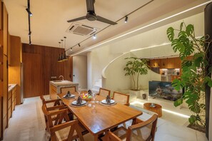 A modern kitchen with wooden cabinets, a dining area with a wooden table and chairs, and a large TV screen. - Lumea Canggu by Pertama Stay (Canggu)