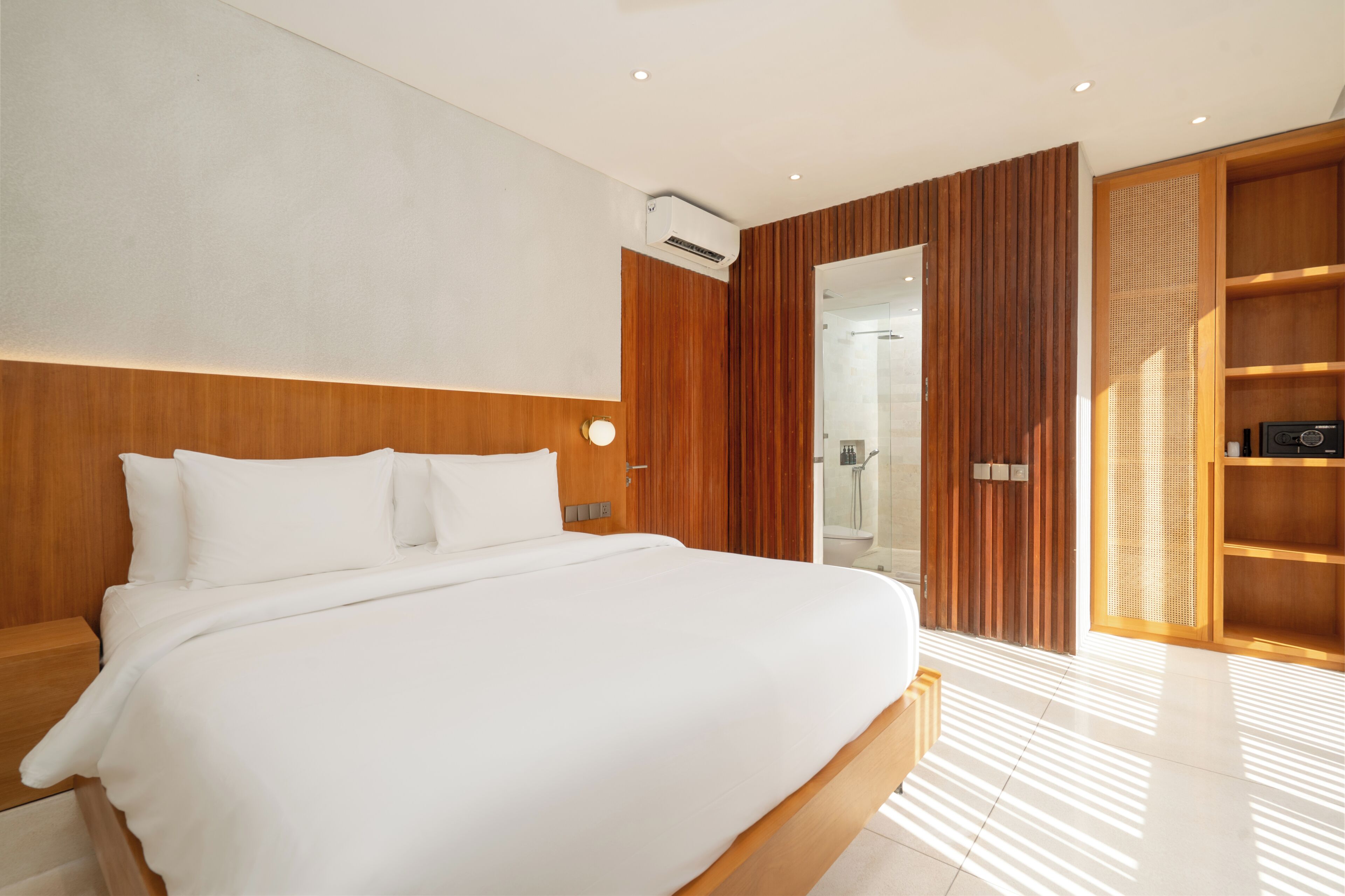 A modern hotel room with a large bed, wooden headboard, a walk-in closet, and a bathroom with a glass shower enclosure.