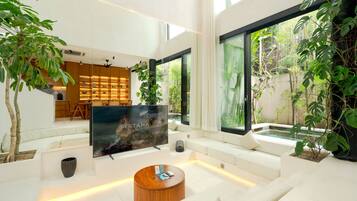Una sala de estar moderna con un televisor de pantalla plana, sofás blancos, una mesa de centro y amplios ventanales con vistas a un jardín.