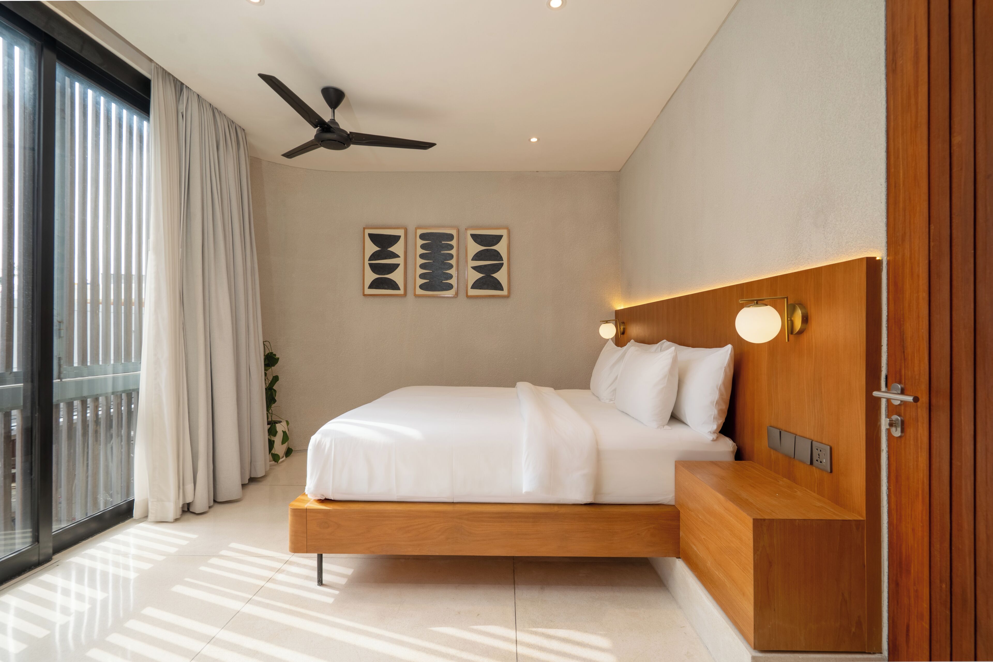 A modern bedroom with a bed, ceiling fan, wall art, bedside lamp, and a view through a large window.