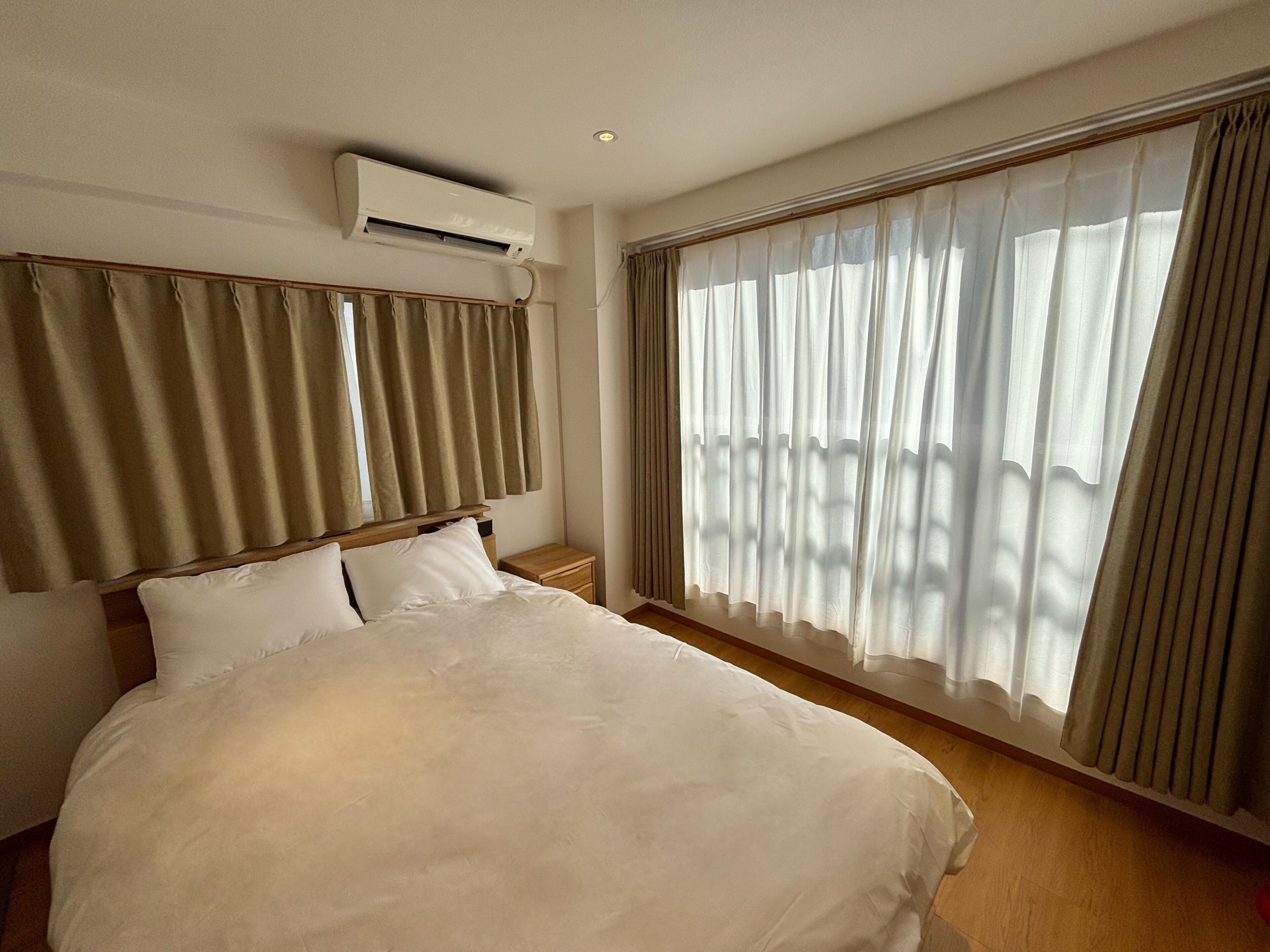 A hotel room with a large bed, an air conditioning unit, a wooden headboard, and sheer curtains on the windows.