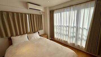 A hotel room with a large bed, an air conditioning unit, a wooden headboard, and sheer curtains on the windows.