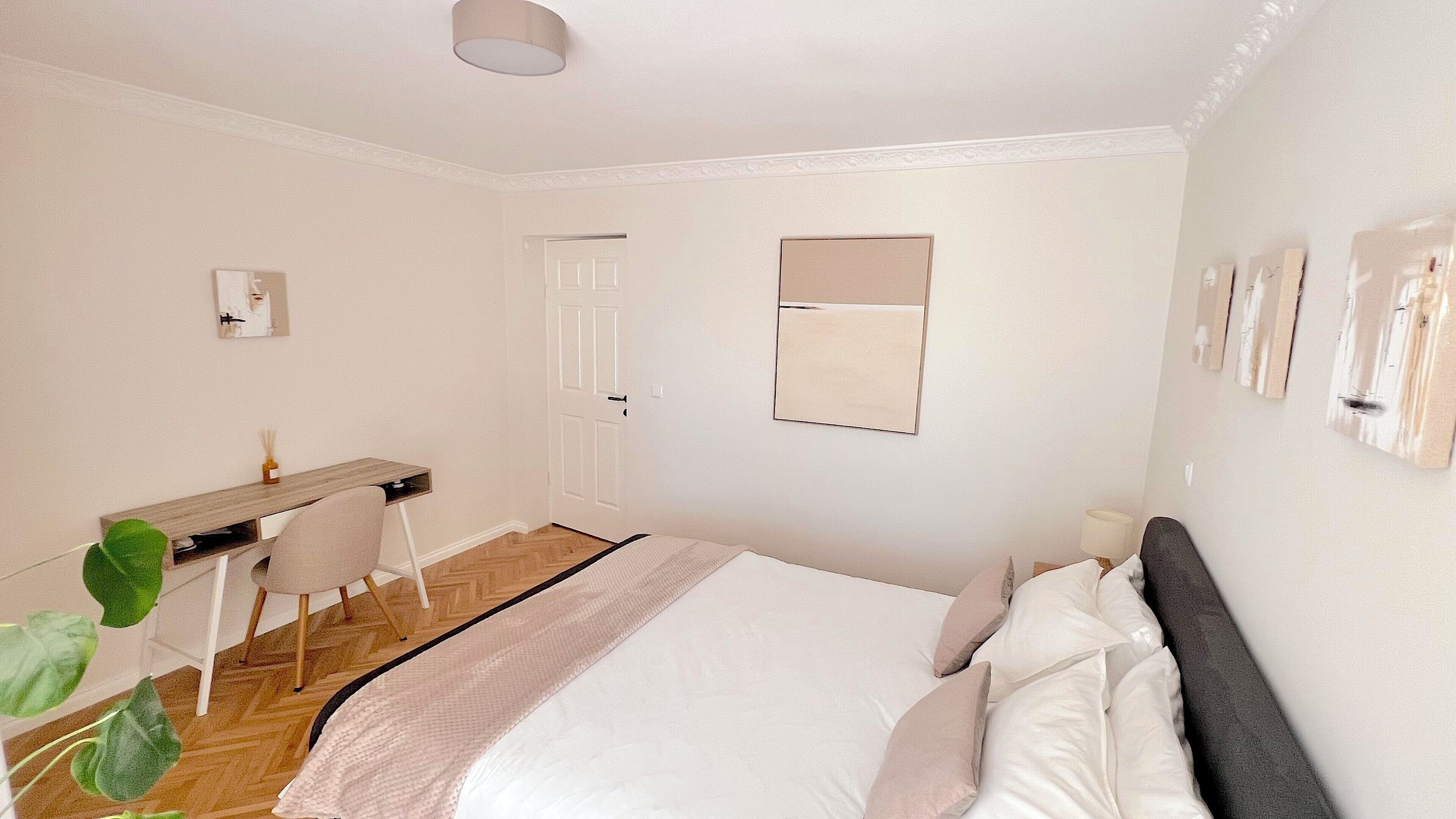 A bedroom with a bed, bedside table, chair, desk, and a mirror on the wall.