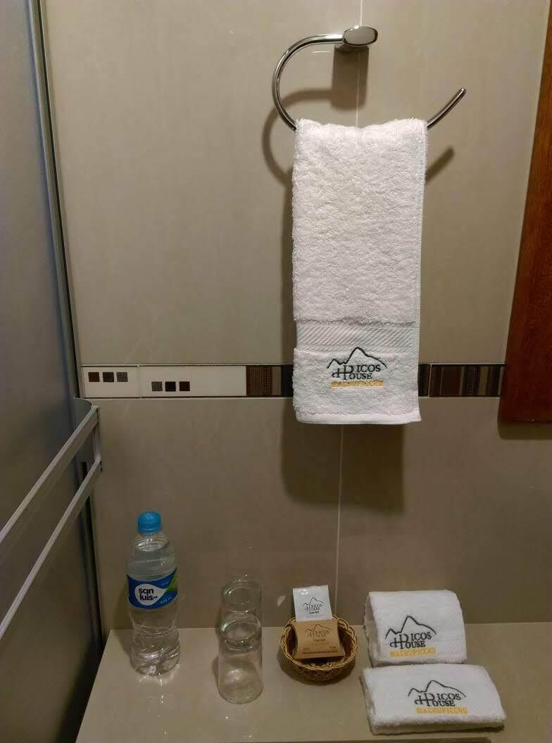 A bathroom with a towel hanging on a metal rack, a bottle of water, two glasses, a soap bar, and two folded towels with a logo.
