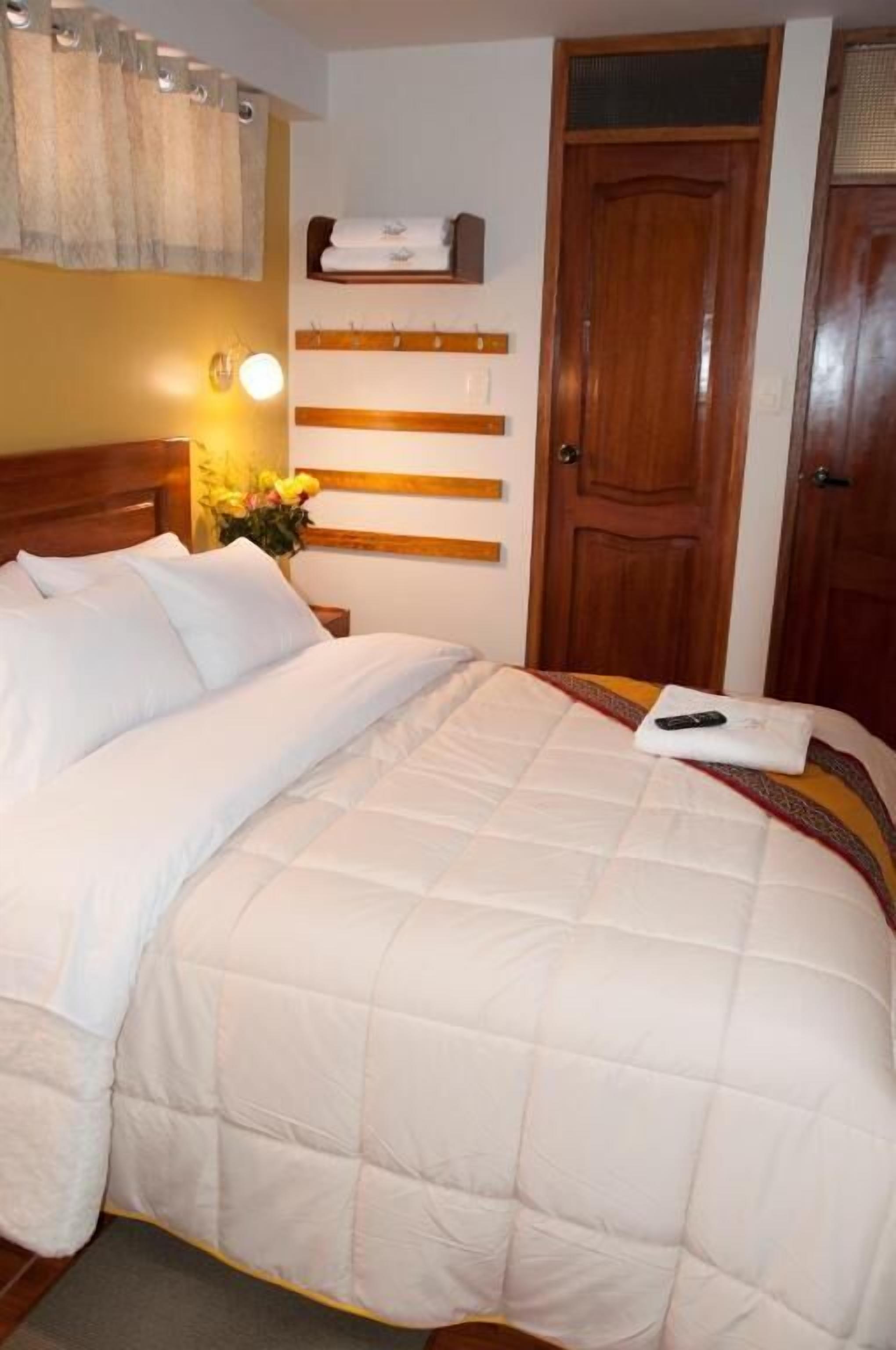 Una cama bien tendida con ropa blanca, un cabecero de madera, un estante en la pared con toallas y una puerta de madera.