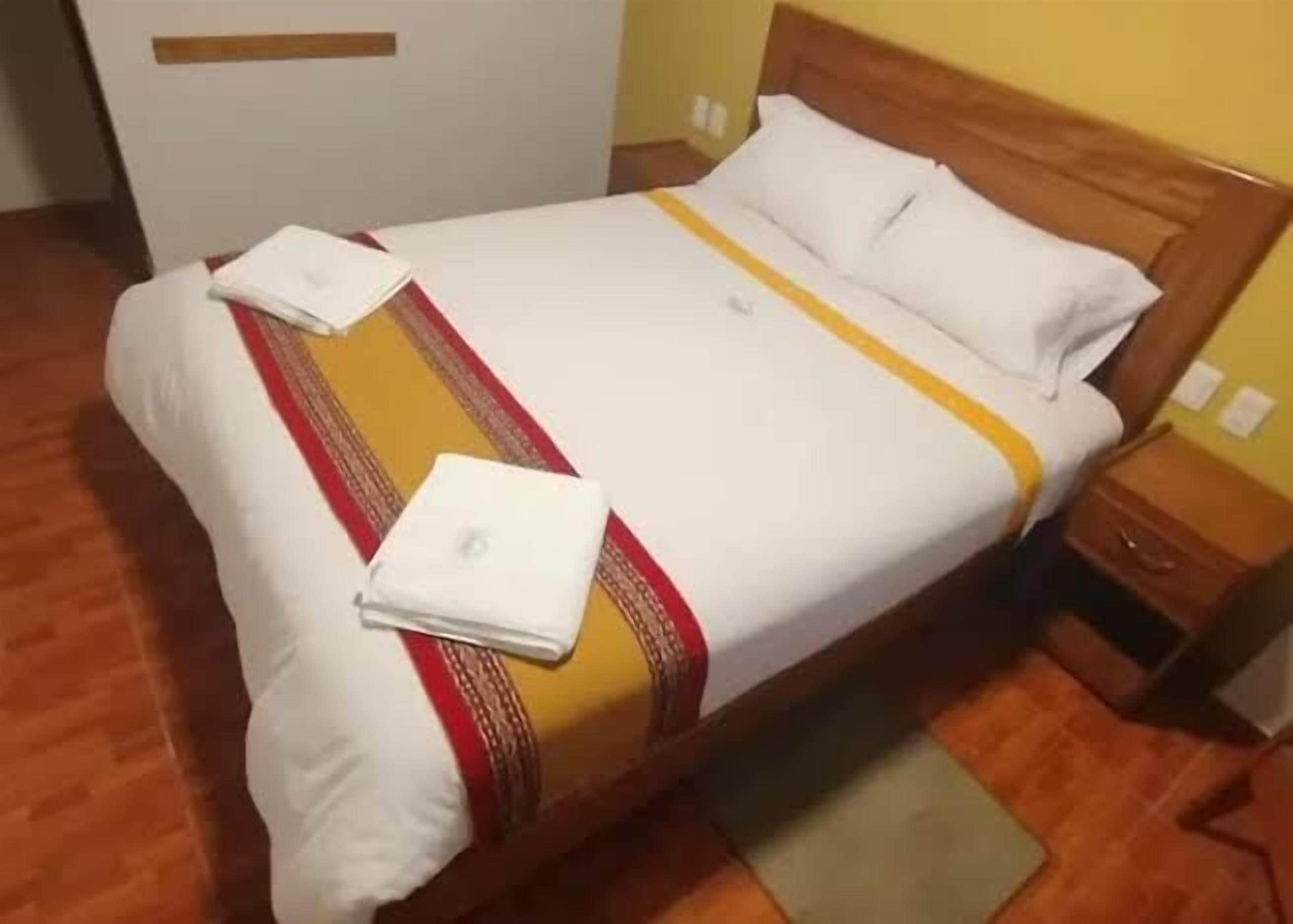 Uma cama bem arrumada com colcha branca e amarela, cabeceira de madeira e uma pequena mesa de cabeceira com luminária.