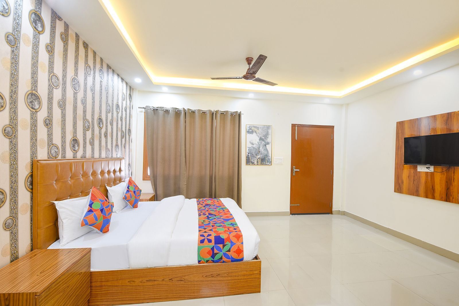 A hotel room with a bed, a wooden headboard, a ceiling fan, a television mounted on the wall, and a door.
