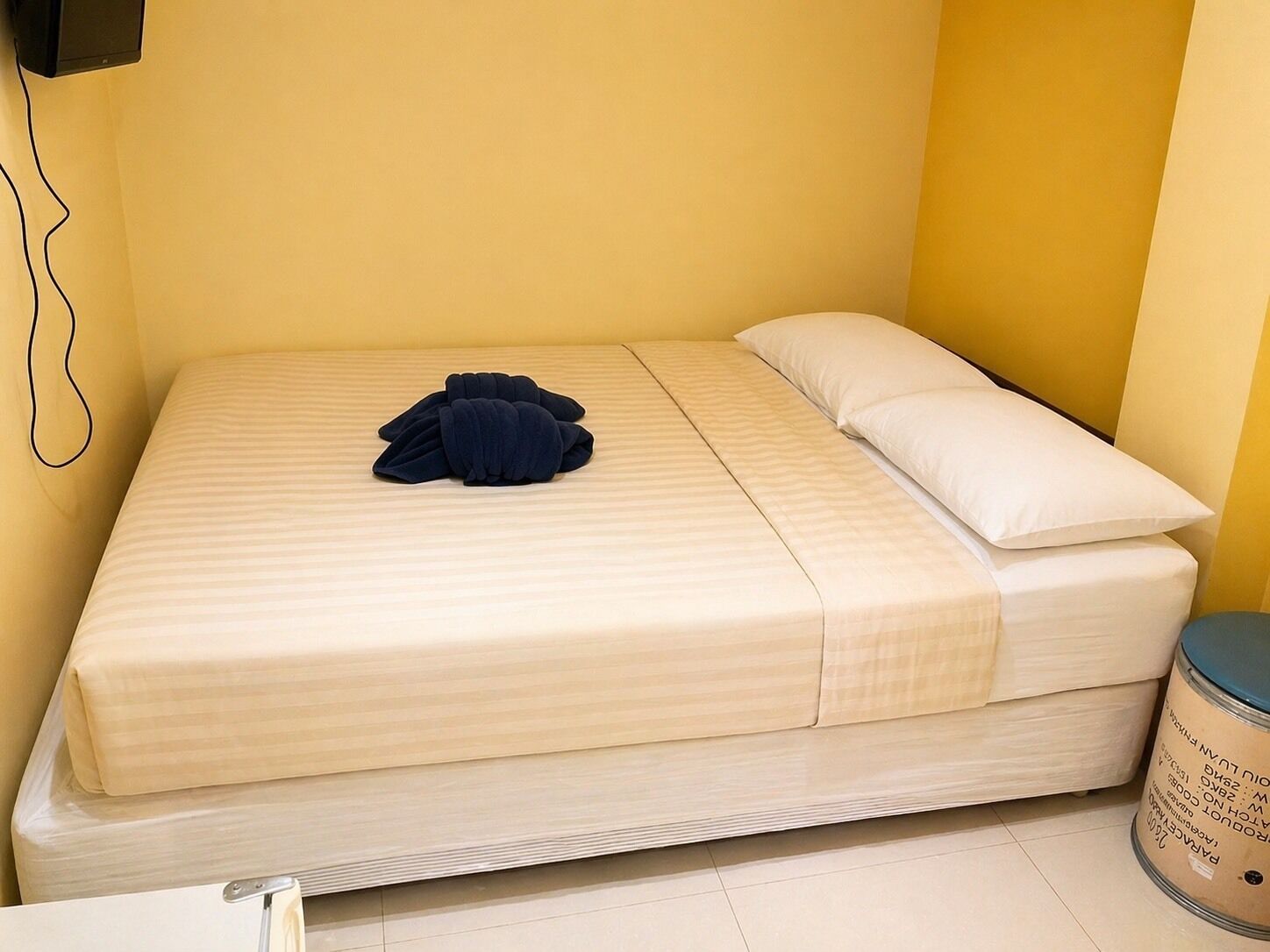 A single bed with a striped mattress, a folded dark blue blanket, and a white pillow in a room with yellow walls and a tiled floor.