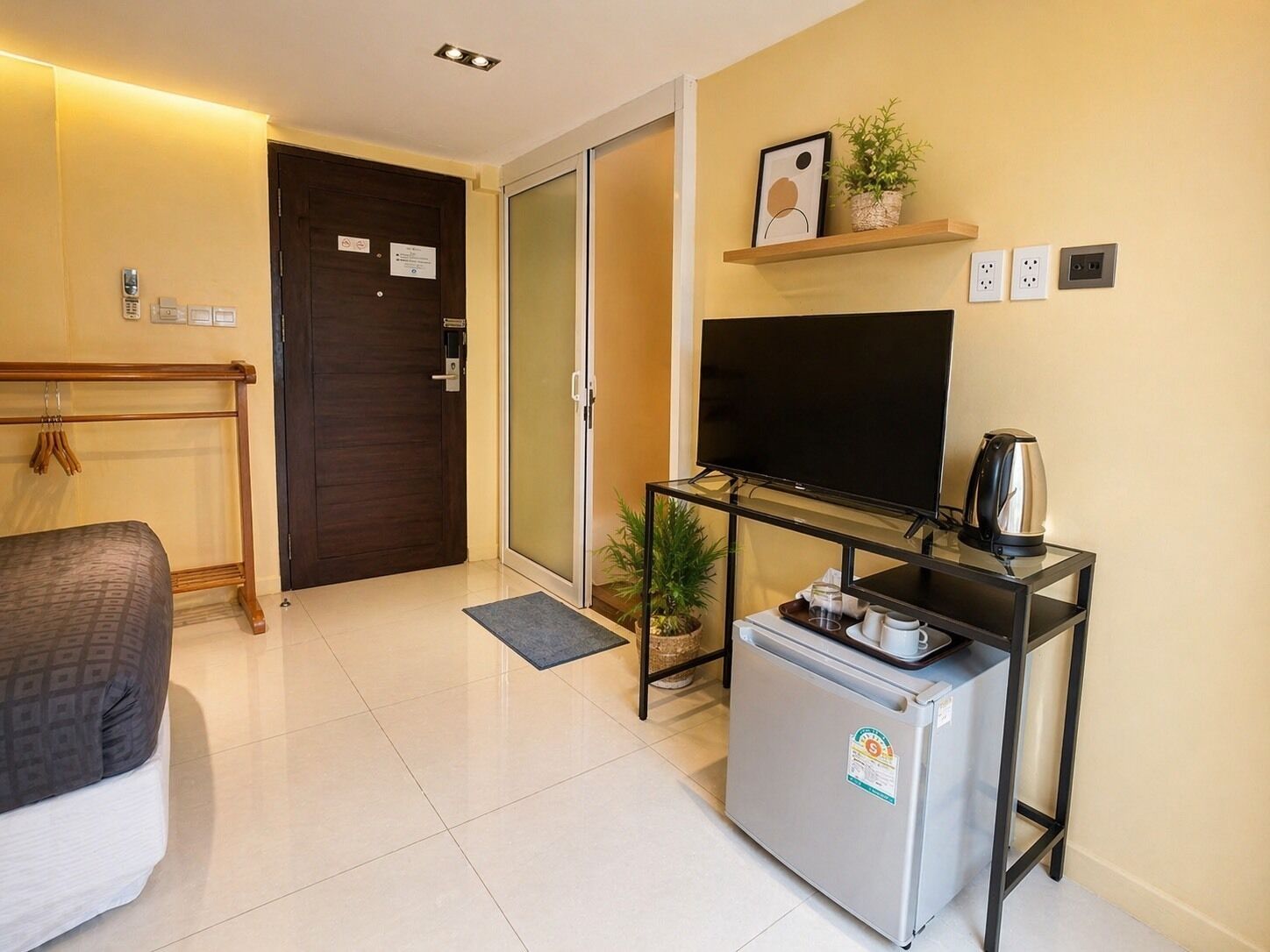 A compact hotel room with a bed, a television, a small refrigerator, a coffee maker, a plant, and a door with a sliding glass panel.