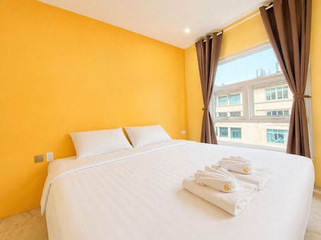 A hotel room with a large bed, white bedding, and a view of buildings through a window with brown curtains.