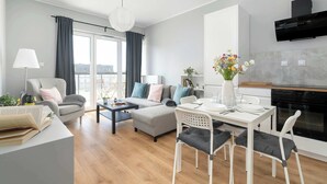 A modern living room with a sofa, dining table, and chairs, a kitchen area with a stove, and a large window with curtains. - Bright Apartment | Parking | Balcony | Remote Work (Poznań)