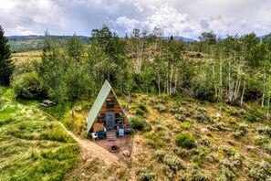 Una cabaña de madera triangular con un porche, rodeada por un paisaje boscoso.