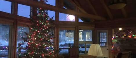 A cozy room with a large window, a Christmas tree, a lamp, and a view of a snowy landscape.