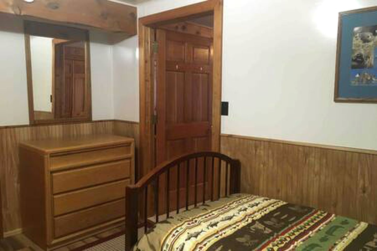 Une chambre avec une commode en bois, un lit recouvert d’une couvre-lit à motifs, une porte en bois et un tableau encadré accroché au mur.