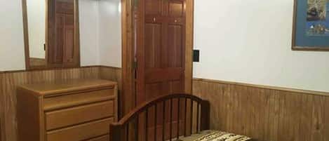 Une chambre avec une commode en bois, un lit recouvert d’une couvre-lit à motifs, une porte en bois et un tableau encadré accroché au mur.