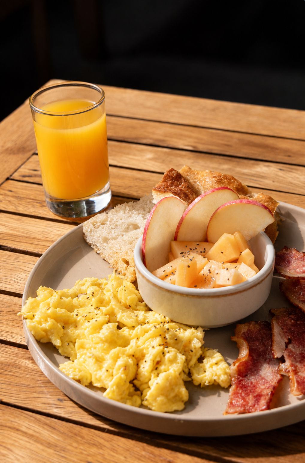 A breakfast plate with scrambled eggs, bacon, toast, apples, and a glass of orange juice on a wooden table.