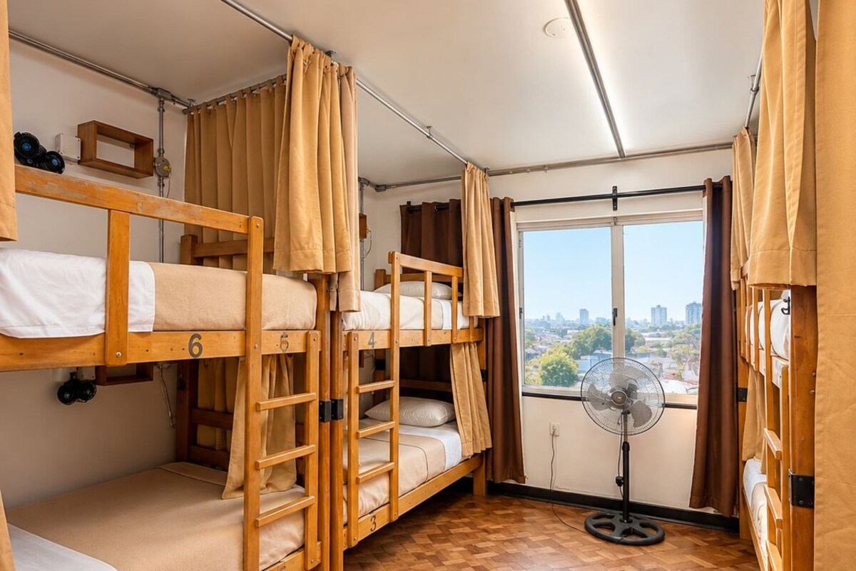 A dormitory room with bunk beds, a fan, and a window with a city view.