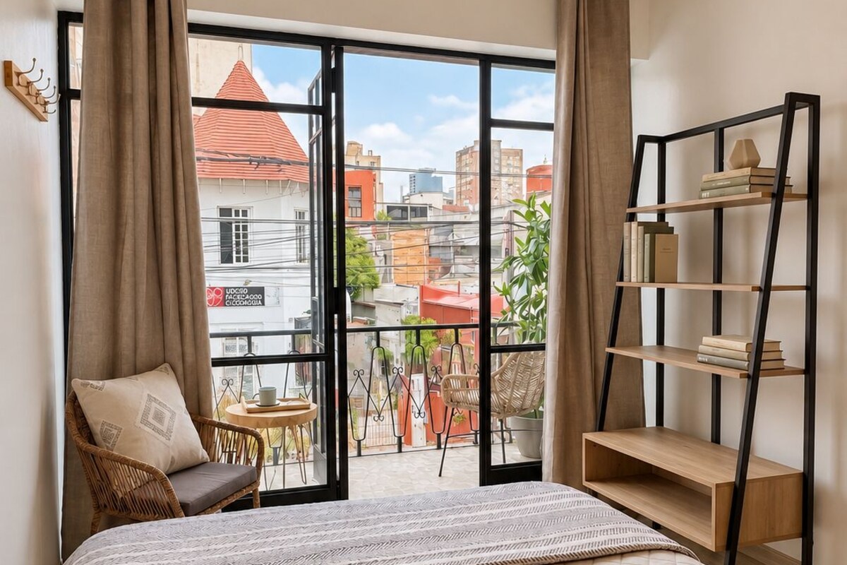 A bedroom with a bed, a bookshelf, a chair, and a balcony with a view of buildings.