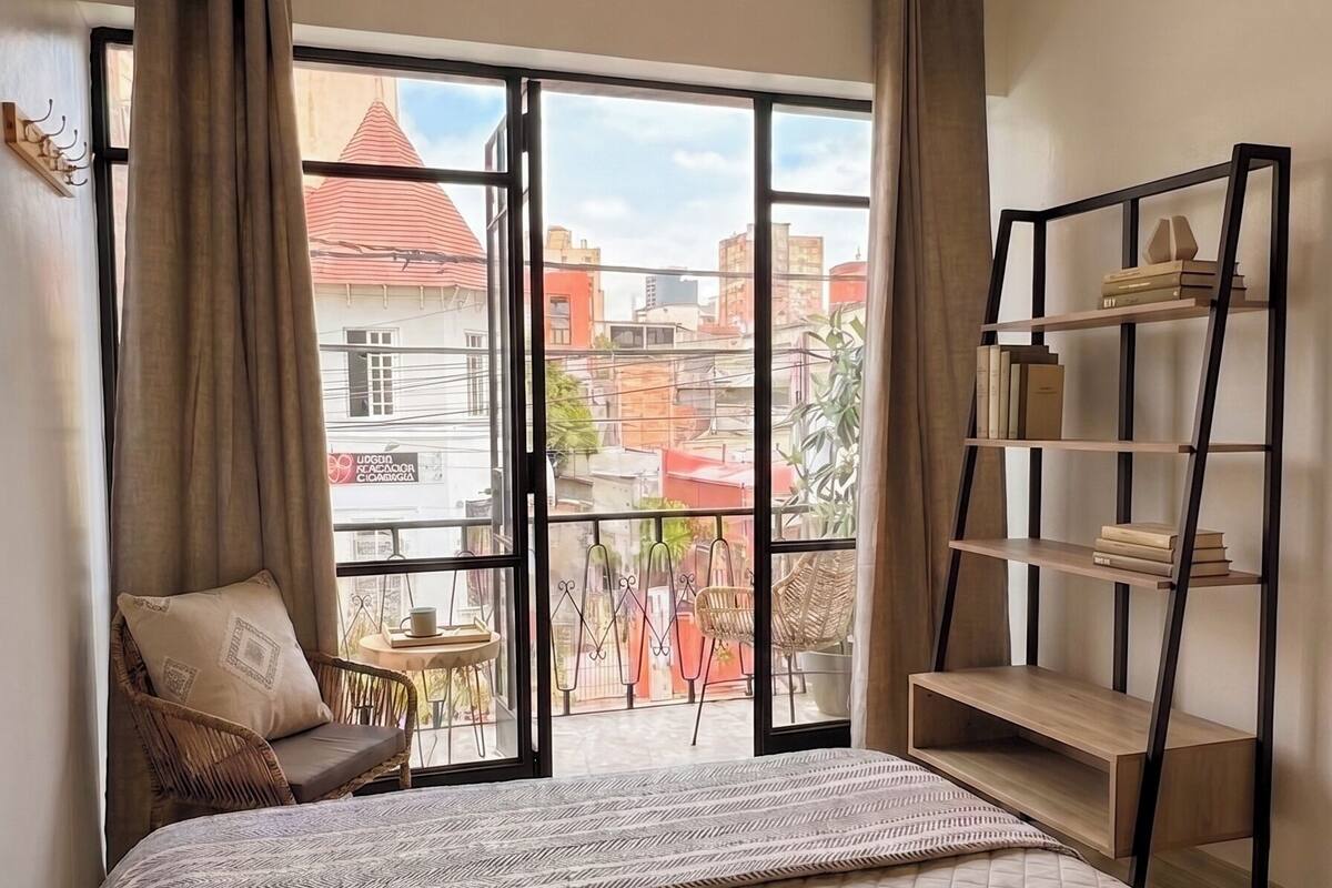A bedroom with a bed, a ceiling fan, a bookshelf, and a view of the city through large windows.