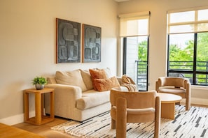 Ein Wohnzimmer mit einer Couch, zwei Sesseln, einem kleinen Beistelltisch und einem großen Fenster mit Blick auf Grünflächen.