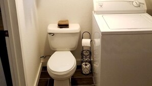 A small, clean bathroom with a toilet, a window, a wall-mounted electrical panel, and a washing machine.