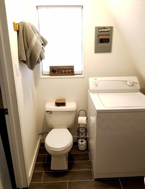 A small, clean bathroom with a toilet, a window, a wall-mounted electrical panel, and a washing machine.