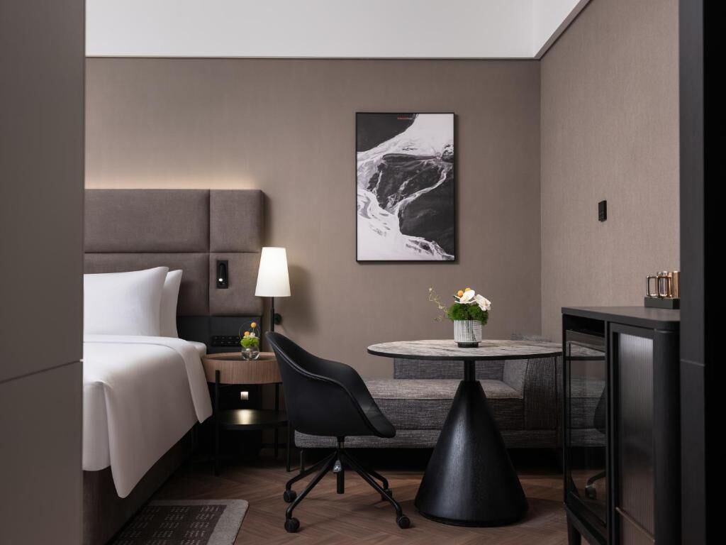 A hotel room with a bed, a desk, a chair, a round table, a lamp, a painting, and a minibar.