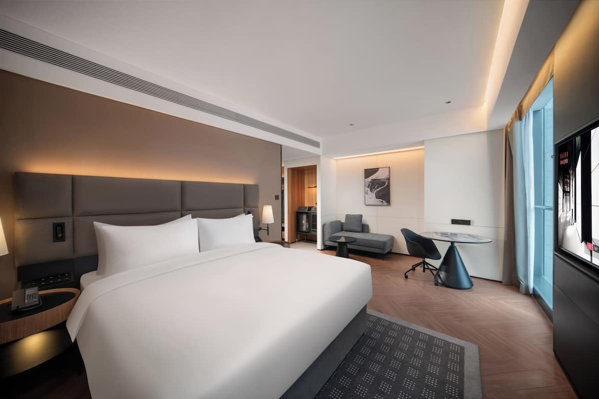 A modern hotel room with a large bed, bedside table with a phone, a desk with a chair, a small table, and a large window.