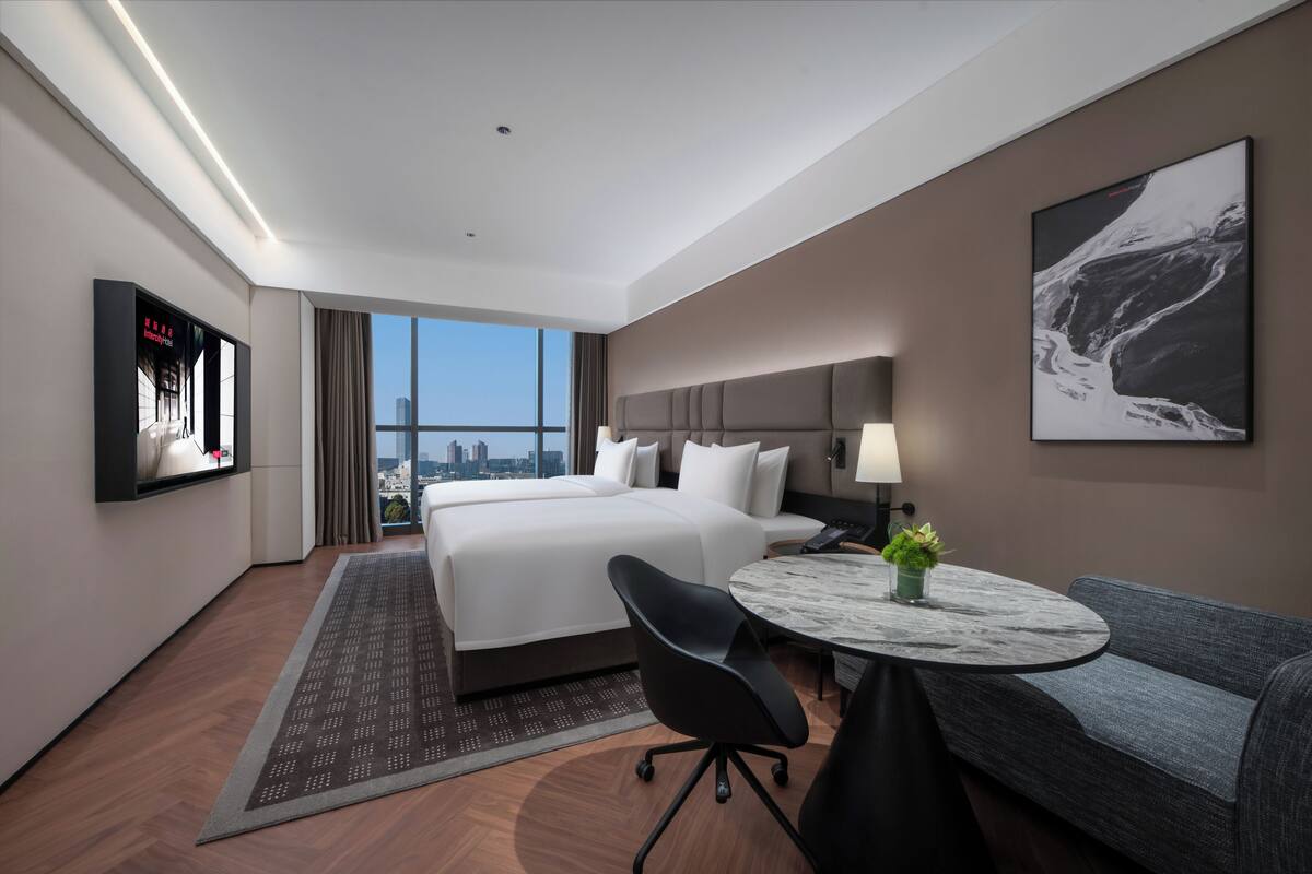 A hotel room with a large bed, a desk with a chair, a sofa, a TV mounted on the wall, and a city view.