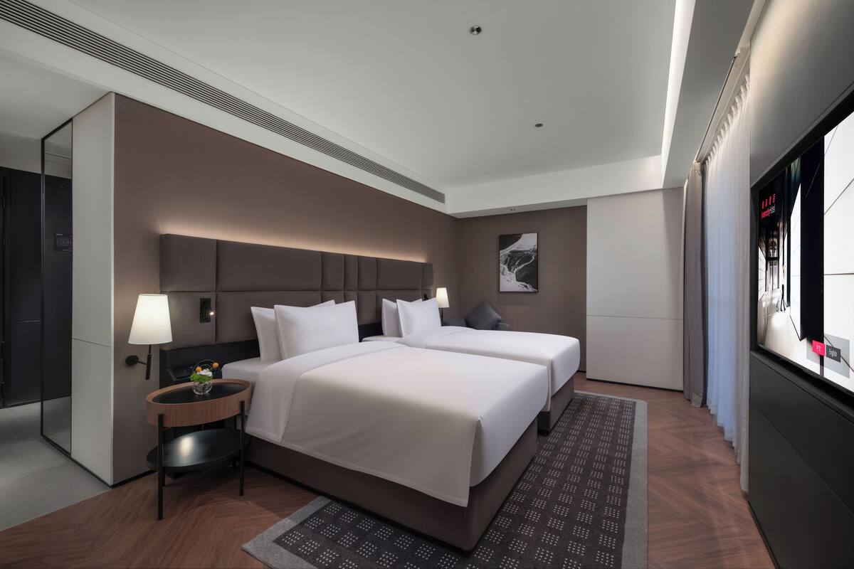 A hotel room with two beds, a bedside table with a lamp, a small plant, a large window, and a flat-screen TV.