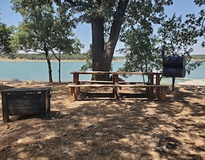 Ein Picknicktisch unter einem Baum am Seeufer, mit Grill und Mülleimer.
