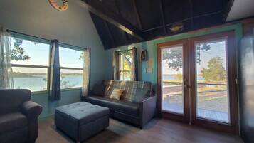 Ein Wohnzimmer mit Sofa, Hocker und Sessel, großen Fenstern mit Jalousien und Blick auf ein Gewässer.