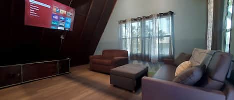 Una sala de estar con un televisor mostrando la interfaz de un servicio de streaming, un sofá marrón, un reposapiés gris y una ventana con cortinas.