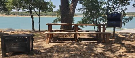 A picnic table under a tree by a lake with a grill and a trash can.
