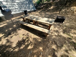 A wooden picnic table in a shaded outdoor area with a metal grill and a black speaker on the ground.