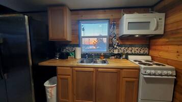 A compact kitchen with wooden cabinets, a stainless steel refrigerator, a microwave, a stove, and a window with blinds.