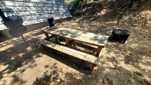 Un tavolo da picnic in legno in una zona esterna ombreggiata, con una griglia in metallo e un altoparlante nero a terra.